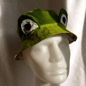 Bob court en sergé vert moyen assez lumineux, et des yeux de grenouille (tour vert, centre blanc avec la pupille noire) dressés au-dessus du rebord en tissu imprimé, fond vert vif imprimé de motifs floraux stylisés et symétriques vert-jaunes Vu de face sur un mannequin de couture