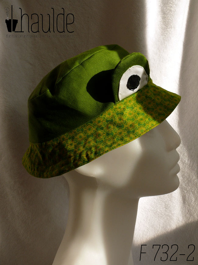 Bob court en sergé vert moyen assez lumineux, et des yeux de grenouille (tour vert, centre blanc avec la pupille noire) dressés au-dessus du rebord en tissu imprimé, fond vert vif imprimé de motifs floraux stylisés et symétriques vert-jaunes Vu de profil sur un mannequin de couture