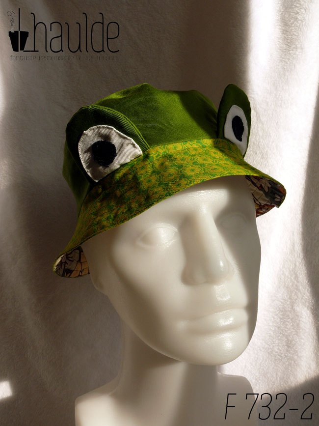 Bob court en sergé vert moyen assez lumineux, et des yeux de grenouille (tour vert, centre blanc avec la pupille noire) dressés au-dessus du rebord en tissu imprimé, fond vert vif imprimé de motifs floraux stylisés et symétriques vert-jaunes Vu de face sur un mannequin de couture