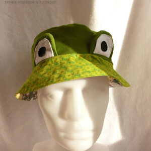 Bob court en sergé vert moyen assez lumineux, et des yeux de grenouille (tour vert, centre blanc avec la pupille noire) dressés au-dessus du rebord en tissu imprimé, fond vert vif imprimé de motifs floraux stylisés et symétriques vert-jaunes Vu de face sur un mannequin de couture