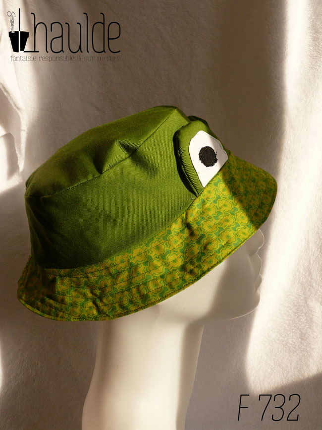 Bob court en sergé vert moyen assez lumineux, et des yeux de grenouille (tour vert, centre blanc avec la pupille noire) dressés au-dessus du rebord en tissu imprimé, fond vert vif imprimé de motifs floraux stylisés et symétriques vert-jaunes Vu de profil sur un mannequin de couture