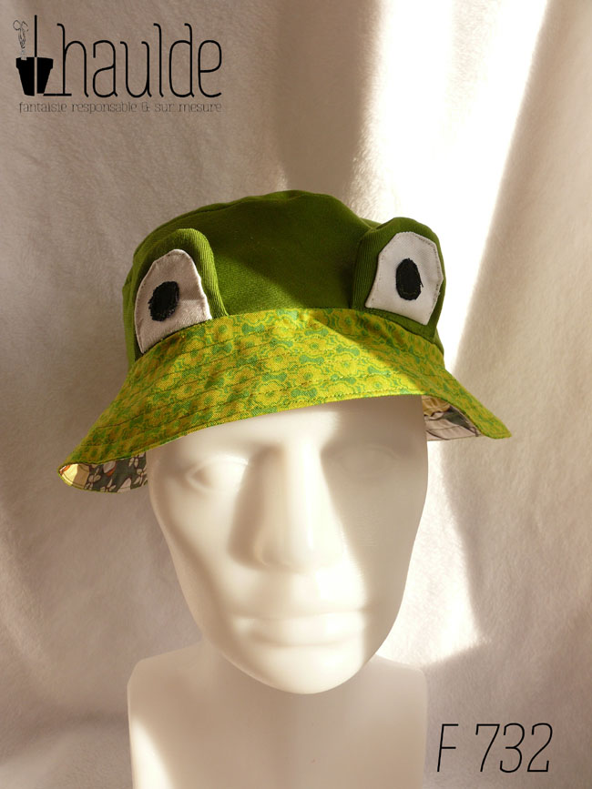 Bob court en sergé vert moyen assez lumineux, et des yeux de grenouille (tour vert, centre blanc avec la pupille noire) dressés au-dessus du rebord en tissu imprimé, fond vert vif imprimé de motifs floraux stylisés et symétriques vert-jaunes Vu de face sur un mannequin de couture