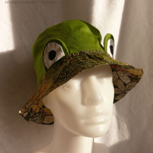 Bob réversible en sergé vert moyen assez lumineux, et des yeux de grenouille (tour vert, centre blanc avec la pupille noire) dressés au-dessus du rebord en tissu imprimé, fond vert kaki foncé comme une aquarelle diluée avec du marron et des ronds vert-jaune dessinés dessus. Vu de trois quarts face sur une tête de mannequin, sous le rebord on devine le tissu de l'envers en tissu kaki imprimé de grandes fleurs jaunes et oranges façon années 70