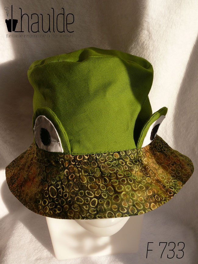 Bob réversible en sergé vert moyen assez lumineux, et des yeux de grenouille (tour vert, centre blanc avec la pupille noire) dressés au-dessus du rebord en tissu imprimé, fond vert kaki foncé comme une aquarelle diluée avec du marron et des ronds vert-jaune dessinés dessus. Vu de dessus