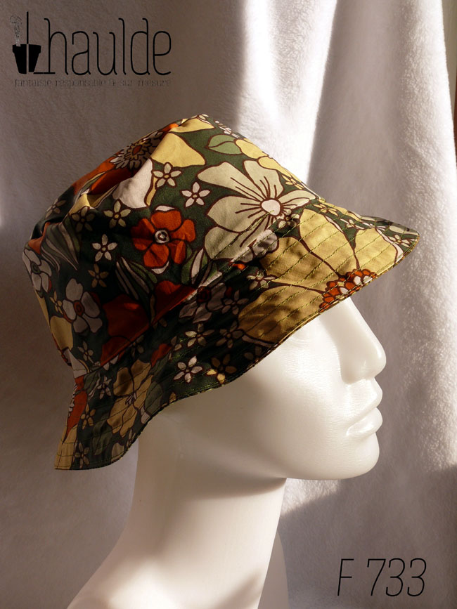 Bob réversible, vu du côté en toile enduite, fond kaki imprimé de grandes fleurs oranges et jaune pâle dans le style des années 70. Vu de profil sur une tête de mannequin