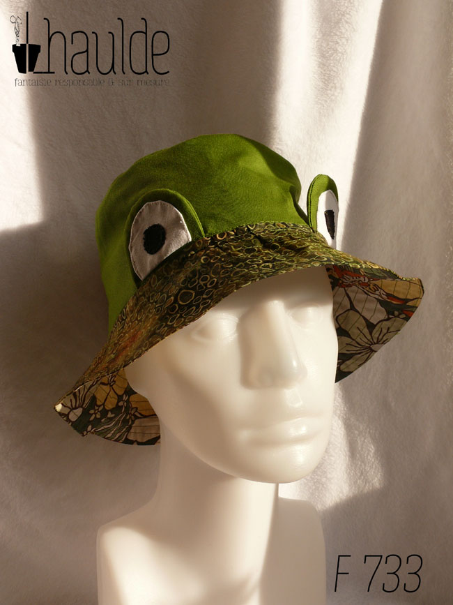 Bob réversible en sergé vert moyen assez lumineux, et des yeux de grenouille (tour vert, centre blanc avec la pupille noire) dressés au-dessus du rebord en tissu imprimé, fond vert kaki foncé comme une aquarelle diluée avec du marron et des ronds vert-jaune dessinés dessus. Vu de trois quarts face sur une tête de mannequin, sous le rebord on devine le tissu de l'envers en tissu kaki imprimé de grandes fleurs jaunes et oranges façon années 70