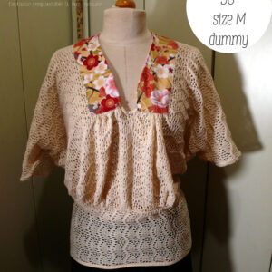 Haut manches mi longues en tricot de coton écru très ajouré, façon crochet, à motifs queues de paons en lignes horizontales. Le col plat est en tissu imprimé, fond doré avec de grosses fleurs de cerisier rouges, blanches et roses, soulignées de vrai doré. Le haut est ample et froncé jusqu'au hanches qu'il moule. Les manches descendent jusqu'au niveau du coude dessus, mais sont plus courtes dessous Vu de face sur un mannequin de couture réglé en taille 38/M