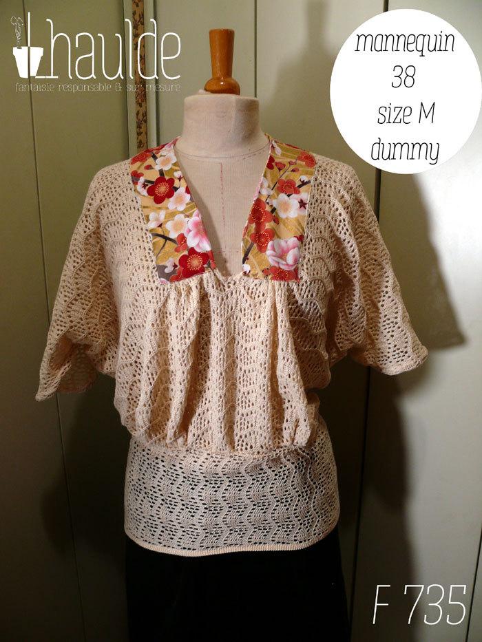 Haut manches mi longues en tricot de coton écru très ajouré, façon crochet, à motifs queues de paons en lignes horizontales. Le col plat est en tissu imprimé, fond doré avec de grosses fleurs de cerisier rouges, blanches et roses, soulignées de vrai doré. Le haut est ample et froncé jusqu'au hanches qu'il moule. Les manches descendent jusqu'au niveau du coude dessus, mais sont plus courtes dessous Vu de face sur un mannequin de couture réglé en taille 38/M