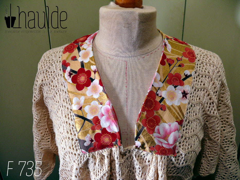 Vue de détail de l'encolure Haut manches mi longues en tricot de coton écru très ajouré, façon crochet, à motifs queues de paons en lignes horizontales. Le col plat est en tissu imprimé, fond doré avec de grosses fleurs de cerisier rouges, blanches et roses, soulignées de vrai doré. Le haut est ample et froncé jusqu'au hanches qu'il moule. Les manches descendent jusqu'au niveau du coude dessus, mais sont plus courtes dessous Vu de face sur un mannequin de couture réglé en taille 38/M
