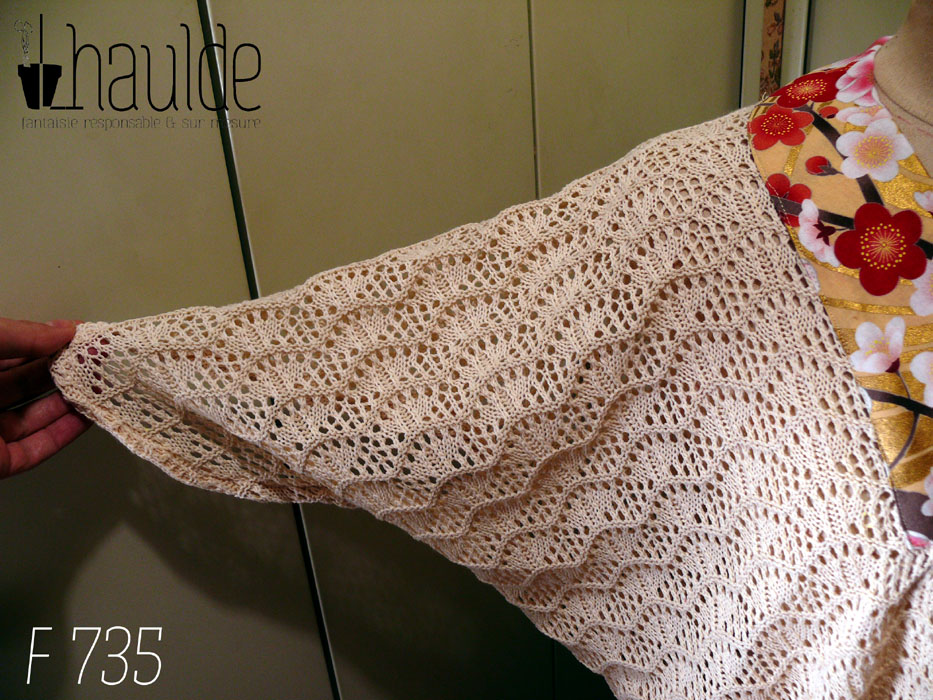 Vue de détail de la manche déployée en longueur, la coupe est assez "chauve-souris" Haut manches mi longues en tricot de coton écru très ajouré, façon crochet, à motifs queues de paons en lignes horizontales. Le col plat est en tissu imprimé, fond doré avec de grosses fleurs de cerisier rouges, blanches et roses, soulignées de vrai doré. Le haut est ample et froncé jusqu'au hanches qu'il moule. Les manches descendent jusqu'au niveau du coude dessus, mais sont plus courtes dessous Vu de face sur un mannequin de couture réglé en taille 38/M
