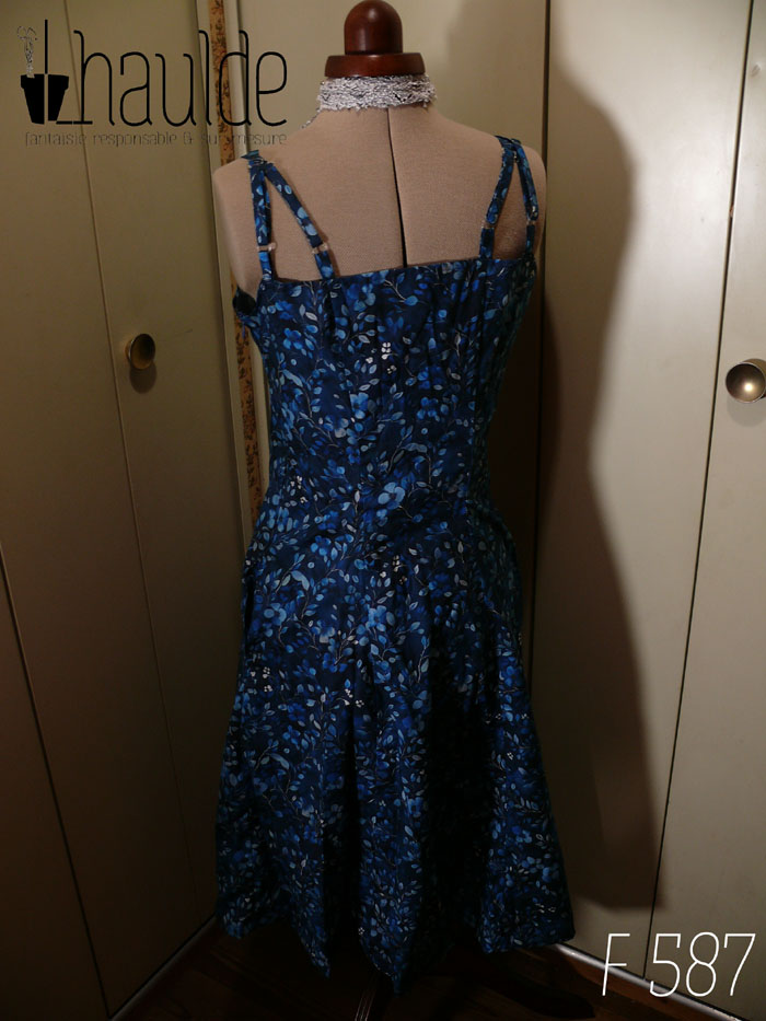 Robe mi-longue avec deux paires de fines bretelles réglables, buste plissé, taille ajustée, jupe ample, en popeline bleu nuit imprimé petite fleurs et végétaux délicats en dégradé de bleu jean. Vue de dos sur un mannequin de couture