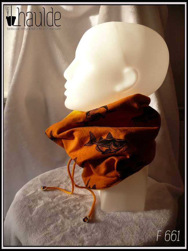 Tour de cou en jersey imprimé et velours ocre, vu côté jersey, fond ocre, motifs carpes noires. Il est porté par une tête de mannequin de profil. Le cordon est également jaune avec des cloches pour cacher les noeuds au bout et un stop pour régler les cordons Il est fourré de polaire de coton
