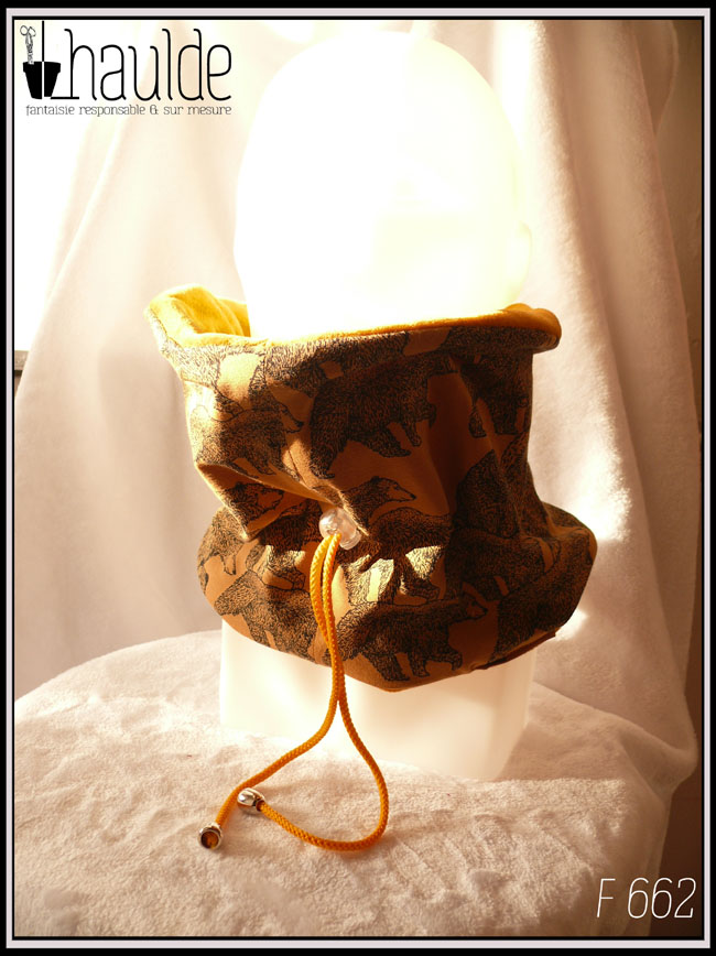 Tour de cou en jersey imprimé, fond ocre, motifs ours de type grizzly en train de marcher ou assis, dessin à la plume. Réversible sur du velours ocre, cordon jaune avec stop, fourré de polaire de coton Vu de trois quarts face sur une tête de mannequin
