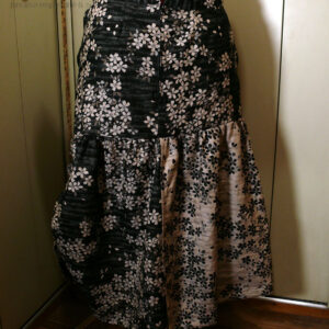 Jupe en coton jacquard, une face noire avec des fleurs de cerisier blanches, une face blanche avec des fleurs de cerisier noires Fond noir pour le haut de la jupe, un pan de chaque couleur pour le bas. Le haut est ajusté, le bas est froncé horizontalement tout le long et verticalement côté gauche, ce qui remonte la jupe sur ce côté là. Deux petits pans de tissu supplémentaires pendent devant, un fond noir sur le fond blanc et inversement de l'autre côté. Vue de dos sur un mannequin de couture. Dos zippé