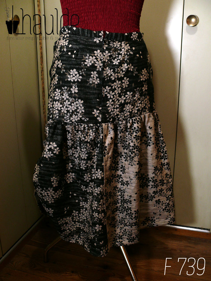 Jupe en coton jacquard, une face noire avec des fleurs de cerisier blanches, une face blanche avec des fleurs de cerisier noires Fond noir pour le haut de la jupe, un pan de chaque couleur pour le bas. Le haut est ajusté, le bas est froncé horizontalement tout le long et verticalement côté gauche, ce qui remonte la jupe sur ce côté là. Deux petits pans de tissu supplémentaires pendent devant, un fond noir sur le fond blanc et inversement de l'autre côté. Vue de dos sur un mannequin de couture. Dos zippé