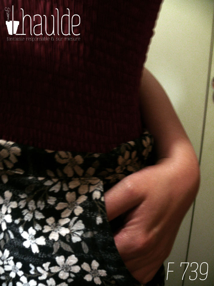 Jupe en coton jacquard, une face noire avec des fleurs de cerisier blanches, une face blanche avec des fleurs de cerisier noires Fond noir pour le haut de la jupe, un pan de chaque couleur pour le bas. Le haut est ajusté, le bas est froncé horizontalement tout le long et verticalement côté gauche, ce qui remonte la jupe sur ce côté là. Deux petits pans de tissu supplémentaires pendent devant, un fond noir sur le fond blanc et inversement de l'autre côté. Vue de détail d'une des deux poches italiennes avec une main dedans