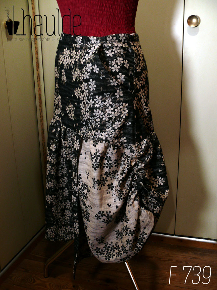 Jupe en coton jacquard, une face noire avec des fleurs de cerisier blanches, une face blanche avec des fleurs de cerisier noires Fond noir pour le haut de la jupe, un pan de chaque couleur pour le bas. Le haut est ajusté, le bas est froncé horizontalement tout le long et verticalement côté gauche, ce qui remonte la jupe sur ce côté là. Deux petits pans de tissu supplémentaires pendent devant, un fond noir sur le fond blanc et inversement de l'autre côté. Vue de profil, côté froncé, sur un mannequin de couture.