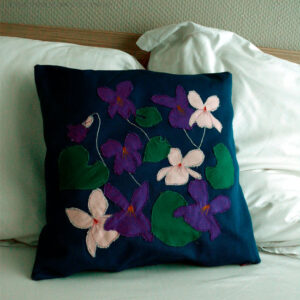 Housse de coussin en sergé bleu roi, avec neuf appliqués en formes de fleurs de violettes, six de couleur violette dont une en bouton et trois roses pâles et cinq appliqués en sergé vert émeraude en forme de feuilles de violettes. Les tiges des fleurs sont parfois représentées par une ligne de point brodé à la machine. Vu de face sur un lit blanc devant deux oreillers blancs.