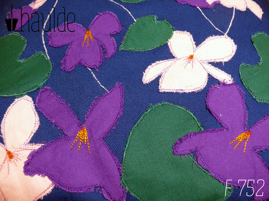 Housse de coussin en sergé bleu roi, avec neuf appliqués en formes de fleurs de violettes, six de couleur violette dont une en bouton et trois roses pâles et cinq appliqués en sergé vert émeraude en forme de feuilles de violettes. Les tiges des fleurs sont parfois représentée par une ligne de point brodé à la machine. Vue en gros plan