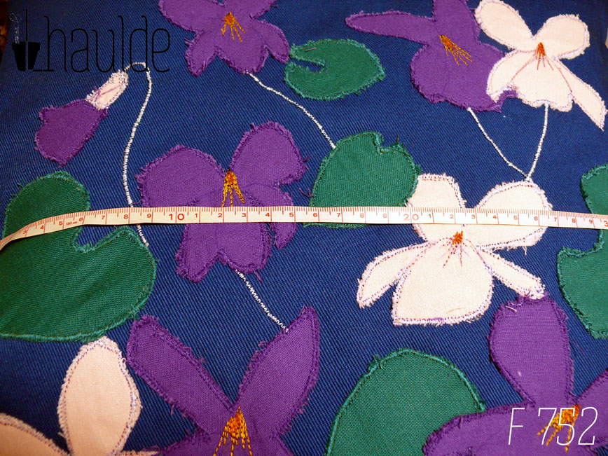 Housse de coussin en sergé bleu roi, avec neuf appliqués en formes de fleurs de violettes, six de couleur violette dont une en bouton et trois roses pâles et cinq appliqués en sergé vert émeraude en forme de feuilles de violettes. Les tiges des fleurs sont parfois représentée par une ligne de point brodé à la machine. Vu de face en gros plan avec un mètre posé dessus, les motifs fonts de 5 à 8 cm de large