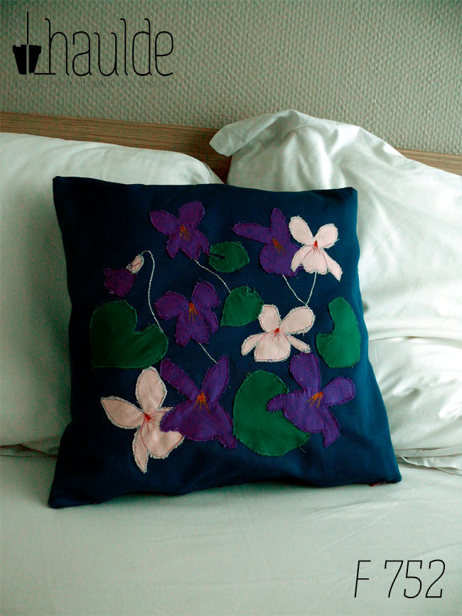 Housse de coussin en sergé bleu roi, avec neuf appliqués en formes de fleurs de violettes, six de couleur violette dont une en bouton et trois roses pâles et cinq appliqués en sergé vert émeraude en forme de feuilles de violettes. Les tiges des fleurs sont parfois représentées par une ligne de point brodé à la machine. Vu de face sur un lit blanc devant deux oreillers blancs.