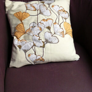 Housse de coussin blanc cassé avec des appliqués en tissu imprimé feuilles de ginkgo translucide et d'appliqués en forme de feuille de ginkgo en sergé jaune orangé surpiqué pour faire les nervures Vu sur un fauteuil violet