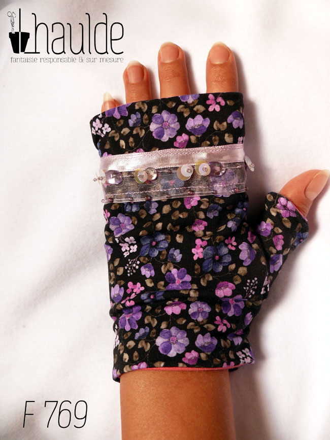 Mitaines en jersey noir imprimé de petites fleurs violettes, le pouce est couvert et au niveau des phalanges les mitaines sont garnies d'un galon d'organza transparent, garni d'un ruban rose pâle et de sequins ronds maintenus par des perles de rocailles Vue d'une mitaine portée, côté dos de la main