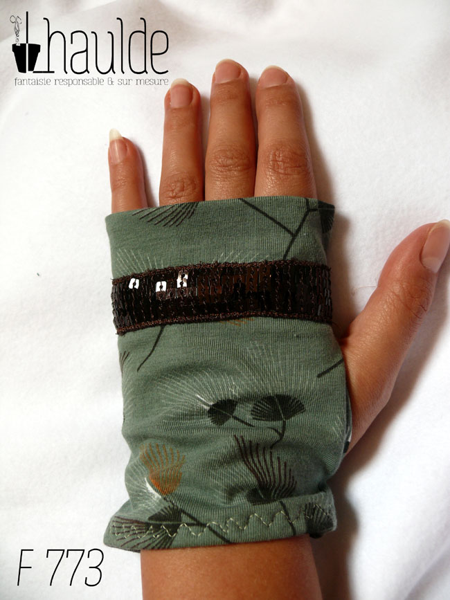 Mitaines en jersey vert céladon imprimé de motifs végétaux en forme de feuilles de palmes stylisées et aérées. Un galon de sequins carrés marrons garnit les mitaines à hauteur des phalanges Vue d'une mitaine portée, côté dos de la main