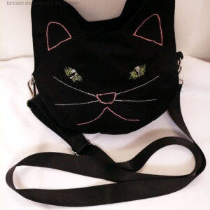 Sac en forme de tête de chat en sergé noir, les oreilles, les yeux, le nez, la moustache et la gueule figurés par des lignes brodées à la machine. Les yeux sont verts, les oreilles, truffe et gueule sont roses, les moustaches bleu pâle Vu posé sur une surface blanche contre un mur blanc, la bandoulière étalée devant. On devine la deuxième paire d'oreilles de l'autre face.