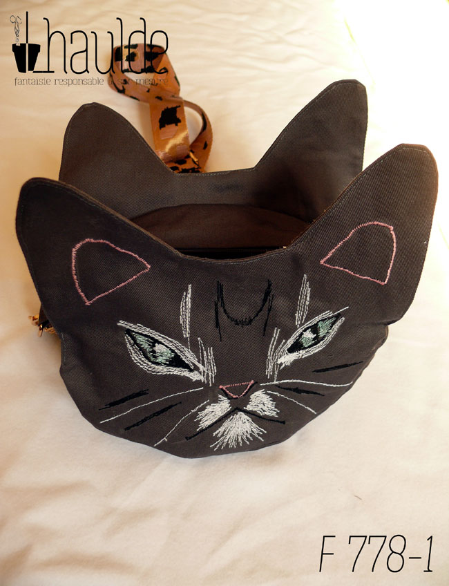 Sac en forme de tête de chat en sergé gris foncé. Les yeux, la moustache et la gueule sont brodés à la machine à coudre de façon assez détaillée, pour représenter les marques d'un chat tigré aux yeux bleus. Les oreilles et la truffe sont figurés par des lignes simples. Les yeux sont entourés de blanc, ainsi que la gueule et des marques noires apparaissent sur le front et les joues. Vu en légère plongée sur une surface blanche, la bandoulière étalée en arrière. Elle est rose pâle avec des motifs léopard noir. On voit la deuxième paire d'oreilles de l'autre face.