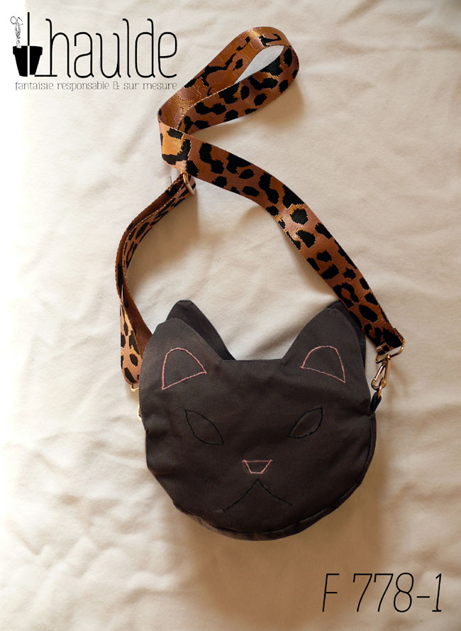 Vue de l'autre face, la tête du chat est à peine esquissée par quelques lignes, oreilles, tour des yeux, truffe et ligne de la gueule Sac en sergé gris foncé avec une bandoulière rose à motifs léopard noirs Vu posé sur une surface blanche