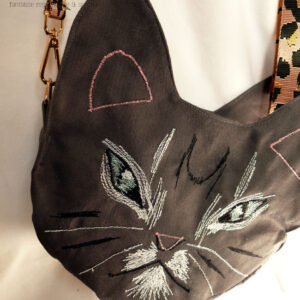 Sac en forme de tête de chat en sergé gris foncé. Les yeux, la moustache et la gueule sont brodés à la machine à coudre de façon assez détaillée, pour représenter les marques d'un chat tigré aux yeux bleus. Les oreilles et la truffe sont figurés par des lignes simples. Les yeux sont entourés de blanc, ainsi que la gueule et des marques noires apparaissent sur le front et les joues. Vu posé sur une surface blanche, la bandoulière étalée en arrière. Elle est rose pâle avec des motifs léopard noir. On devine la deuxième paire d'oreilles de l'autre face. Vu de face, suspendu par sa bandoulière rose à motifs léopard noirs