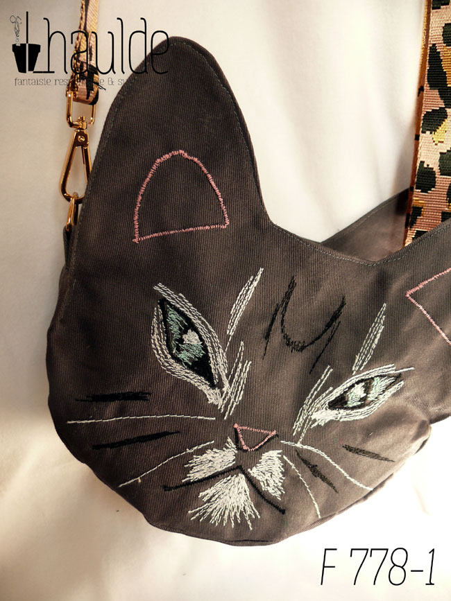 Sac en forme de tête de chat en sergé gris foncé. Les yeux, la moustache et la gueule sont brodés à la machine à coudre de façon assez détaillée, pour représenter les marques d'un chat tigré aux yeux bleus. Les oreilles et la truffe sont figurés par des lignes simples. Les yeux sont entourés de blanc, ainsi que la gueule et des marques noires apparaissent sur le front et les joues. Vu posé sur une surface blanche, la bandoulière étalée en arrière. Elle est rose pâle avec des motifs léopard noir. On devine la deuxième paire d'oreilles de l'autre face. Vu de face, suspendu par sa bandoulière rose à motifs léopard noirs