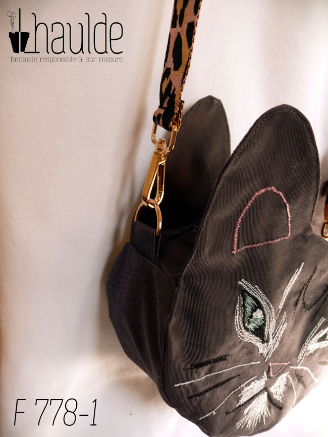 Sac en forme de tête de chat en sergé gris foncé. Les yeux, la moustache et la gueule sont brodés à la machine à coudre de façon assez détaillée, pour représenter les marques d'un chat tigré aux yeux bleus. Les oreilles et la truffe sont figurés par des lignes simples. Les yeux sont entourés de blanc, ainsi que la gueule et des marques noires apparaissent sur le front et les joues. Vu suspendu par sa bandoulière rose à motifs léopard noirs. La tranche du sac avec le mousqueton de la bandoulière sont au centre de l'image
