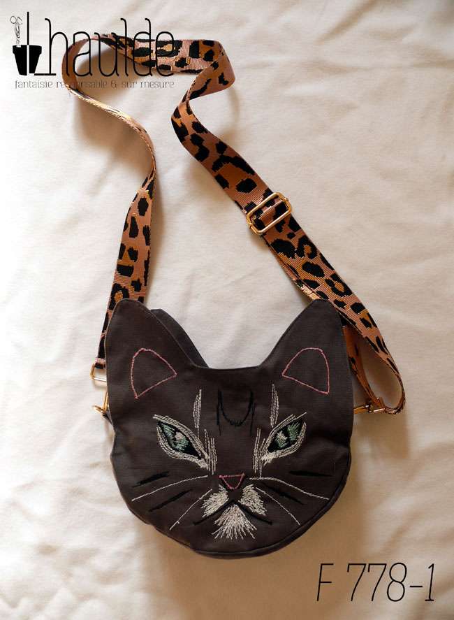 Sac en forme de tête de chat en sergé gris foncé. Les yeux, la moustache et la gueule sont brodés à la machine à coudre de façon assez détaillée, pour représenter les marques d'un chat tigré aux yeux bleus. Les oreilles et la truffe sont figurés par des lignes simples. Les yeux sont entourés de blanc, ainsi que la gueule et des marques noires apparaissent sur le front et les joues. Vu posé sur une surface blanche, la bandoulière étalée en arrière. Elle est rose pâle avec des motifs léopard noir. On devine la deuxième paire d'oreilles de l'autre face.