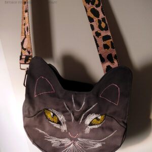 Sac en forme de tête de chat en sergé gris foncé. Les yeux, la moustache et la gueule sont brodés à la machine à coudre de façon assez détaillée, pour représenter les marques d'un chat tigré aux yeux jaunes. Les oreilles et la truffe sont figurés par des lignes simples. Les yeux sont soulignés de blanc, ainsi que la gueule et des marques noires apparaissent sur le front et d'autres descendent en courbe depuis le coin des yeux. Vu suspendu par sa bandoulière, rose pâle avec des motifs léopard noir. On devine la deuxième paire d'oreilles de l'autre face.