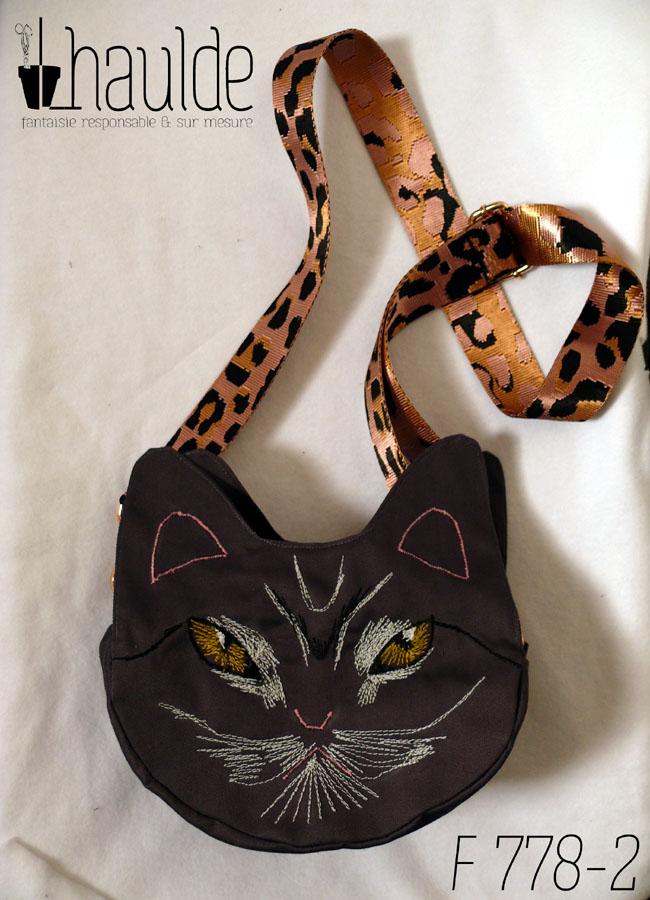 Sac en forme de tête de chat en sergé gris foncé. Les yeux, la moustache et la gueule sont brodés à la machine à coudre de façon assez détaillée, pour représenter les marques d'un chat tigré aux yeux jaunes. Les oreilles et la truffe sont figurés par des lignes simples. Les yeux sont soulignés de blanc, ainsi que la gueule et des marques noires apparaissent sur le front et d'autres descendent en courbe depuis le coin des yeux. Vu posé à plat avec sa bandoulière au-dessus, rose pâle avec des motifs léopard noir. On devine la deuxième paire d'oreilles de l'autre face.