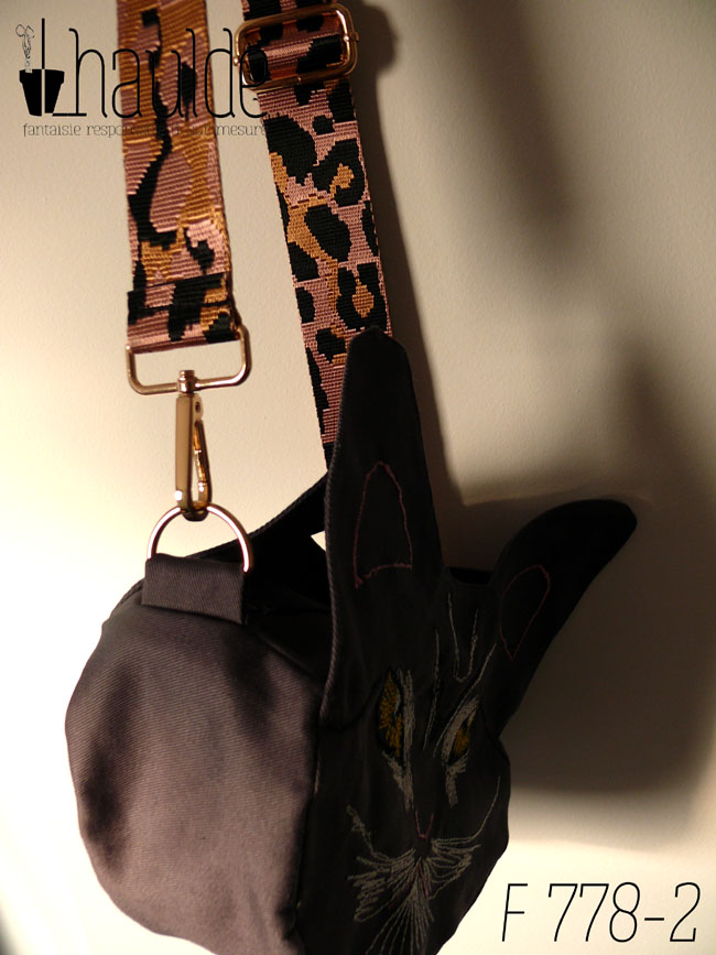 Sac en forme de tête de chat en sergé gris foncé. Les yeux, la moustache et la gueule sont brodés à la machine à coudre de façon assez détaillée, pour représenter les marques d'un chat tigré aux yeux jaunes. Les oreilles et la truffe sont figurés par des lignes simples. Les yeux sont soulignés de blanc, ainsi que la gueule et des marques noires apparaissent sur le front et d'autres descendent en courbe depuis le coin des yeux. Vu suspendu de profil par sa bandoulière, rose pâle avec des motifs léopard noir. On devine la deuxième paire d'oreilles de l'autre face.