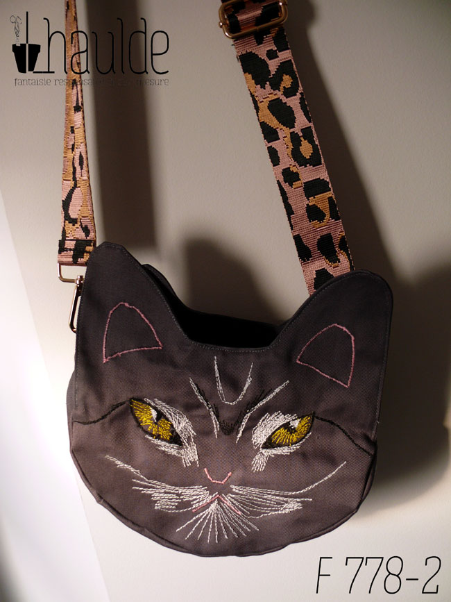 Sac en forme de tête de chat en sergé gris foncé. Les yeux, la moustache et la gueule sont brodés à la machine à coudre de façon assez détaillée, pour représenter les marques d'un chat tigré aux yeux jaunes. Les oreilles et la truffe sont figurés par des lignes simples. Les yeux sont soulignés de blanc, ainsi que la gueule et des marques noires apparaissent sur le front et d'autres descendent en courbe depuis le coin des yeux. Vu suspendu par sa bandoulière, rose pâle avec des motifs léopard noir. On devine la deuxième paire d'oreilles de l'autre face.