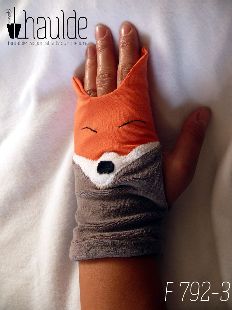 Paire de mitaines en forme de tête de renard. Côté doigts la tête avec les oreilles qui dépassent, en jersey orange, les yeux sont deux lignes noires brodées, le museau noir sur fond blanc est en polaire et le bas de la mitaines est en velours gris. Vue d'une mitaine portée, côté dos de la main