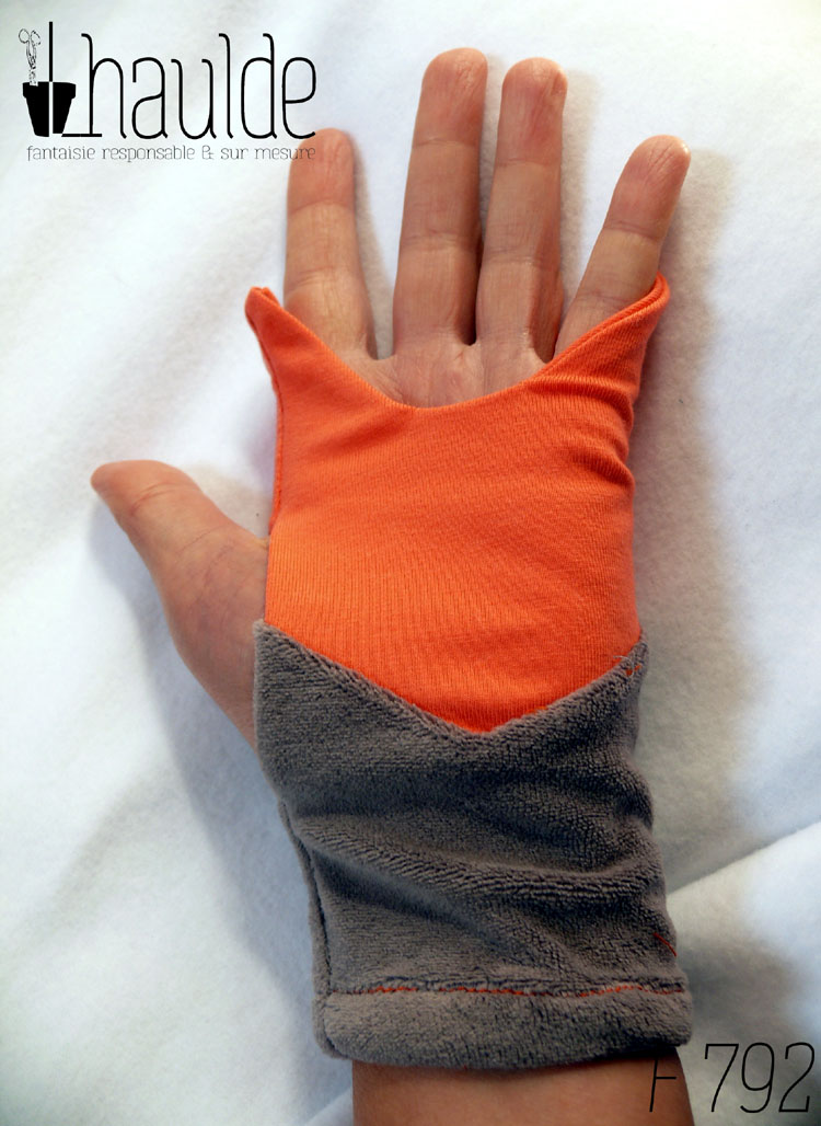 Paire de mitaines en forme de tête de renard. Côté doigts la tête avec les oreilles qui dépassent, en jersey orange, les yeux sont deux lignes noires brodées, le museau noir sur fond blanc est en polaire et le bas de la mitaines est en velours gris. Vue d'une mitaine portée côté paume, il n'y a que le jersey et le velours, pas de dessin de museau
