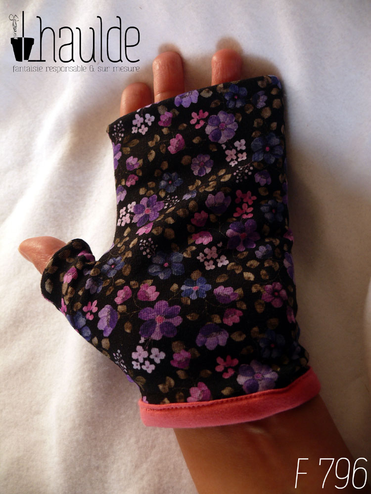 Mitaines avec pouce long en jersey noir imprimé petites fleurs violettes à pétales ronds, par 5 ou par 7 avec des mini fleurs rose très pâle ou rose à 4 pétales et de toutes petites feuilles kakis presque grises Le bord de la manchette est couvert de jersey rose Vue d'une mitaine portée côté paume