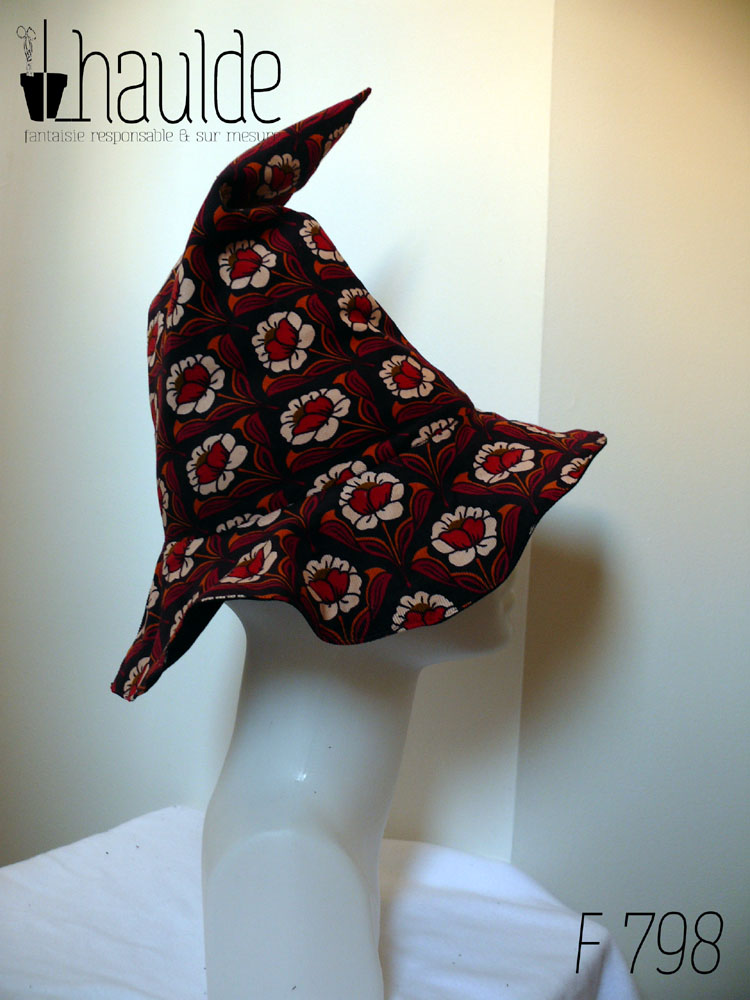 Chapeau de sorcière pointu en velours milleraies marron foncé imprimé de motifs floraux disposés de manière symétrique coeur de la fleur rouge, pétales blancs, feuilles orange et rouge foncé Vu de profil sur une tête de mannequin