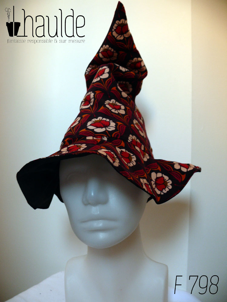 Chapeau de sorcière pointu en velours milleraies marron foncé imprimé de motifs floraux disposés de manière symétrique coeur de la fleur rouge, pétales blancs, feuilles orange et rouge foncé Vu de face sur une tête de mannequin
