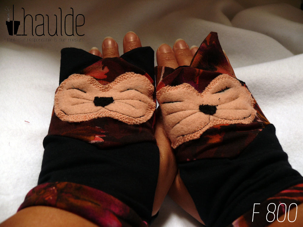 Paire de mitaines en forme de tête de chat, les oreilles dépassent sur les doigts, la tête est dessinée par un tissu polaire rose pale, la truffe en polaire noire, les yeux (fermés, souriants) les moustaches et les babines figurées par une ligne cousue en noir. Le tout cousu sur du jersey imprimé dans les tons rouge bordeaux marron et orange. Le bas de la mitaine est en jersey noir avec une petite manchette rabattue en jersey imprimé. Les oreilles du chat côté gauche sont noires, côté droit sont en tissu imprimé Vue de la paire portée