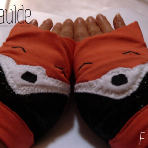 Paire de mitaines en forme de tête de renard. Côté doigts la tête avec les oreilles qui dépassent, en jersey orange, les yeux sont deux lignes noires brodées, le museau noir sur fond blanc est en polaire et le bas de la mitaines est en tissu synthétique noir brillant (comme du velours ras pailleté). Vue de la paire portée