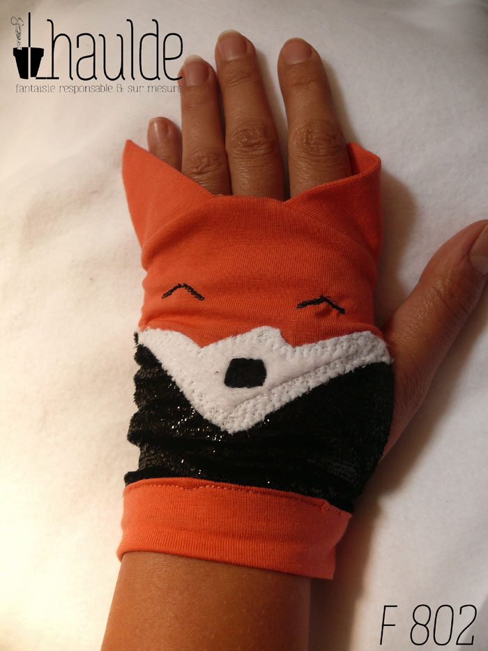 Paire de mitaines en forme de tête de renard. Côté doigts la tête avec les oreilles qui dépassent, en jersey orange, les yeux sont deux lignes noires brodées, le museau noir sur fond blanc est en polaire et le bas de la mitaines est en tissu synthétique noir brillant (comme du velours ras pailleté) Vue d'une mitaine portée vue de dessus