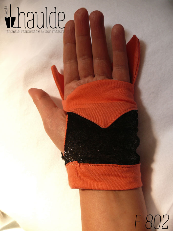 Paire de mitaines en forme de tête de renard. Côté doigts la tête avec les oreilles qui dépassent, en jersey orange, les yeux sont deux lignes noires brodées, le museau noir sur fond blanc est en polaire et le bas de la mitaines est en tissu synthétique noir brillant (comme du velours ras pailleté) Vue d'une mitaine portée, côté paume, le jersey orange monte jusqu'au ras des doigts, on aperçoit les oreilles du renard qui dépassent plus haut, côté doigt.