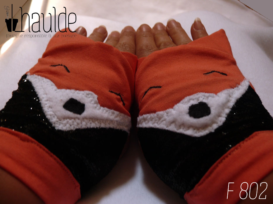 Paire de mitaines en forme de tête de renard. Côté doigts la tête avec les oreilles qui dépassent, en jersey orange, les yeux sont deux lignes noires brodées, le museau noir sur fond blanc est en polaire et le bas de la mitaines est en tissu synthétique noir brillant (comme du velours ras pailleté). Vue de la paire portée